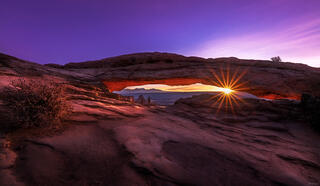 sunburst rising through an arch with purple sky in Canyonlands National Park, Utah