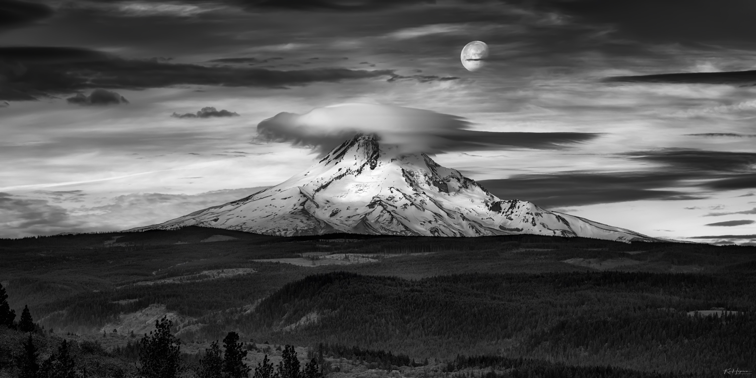 back and white image of mt hood with lenticular cloud capping the mountain with a full moon above, dark clouds and forest surrounding the mountain 