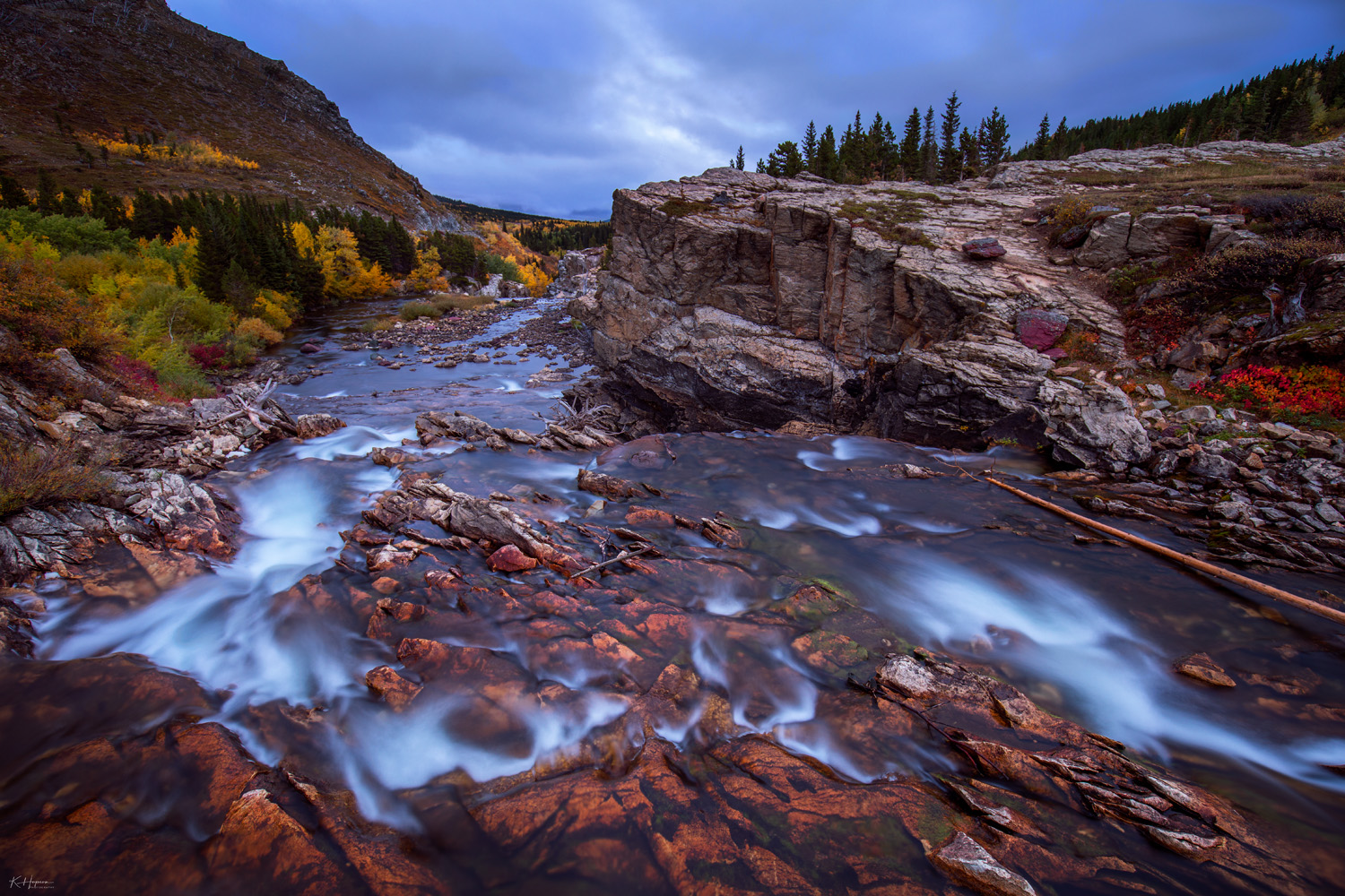 small rapids on a river in Glacier National Park with warm fall colored vegetation on the shoreline with stormy clouds above